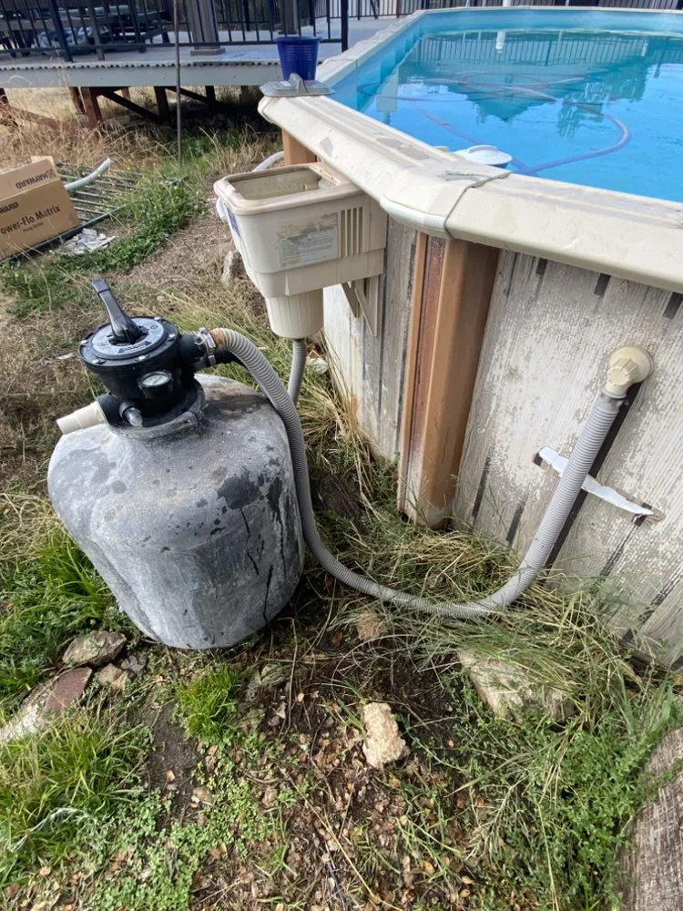 An old sand filter beside an above-ground pool, with weeds around the base
